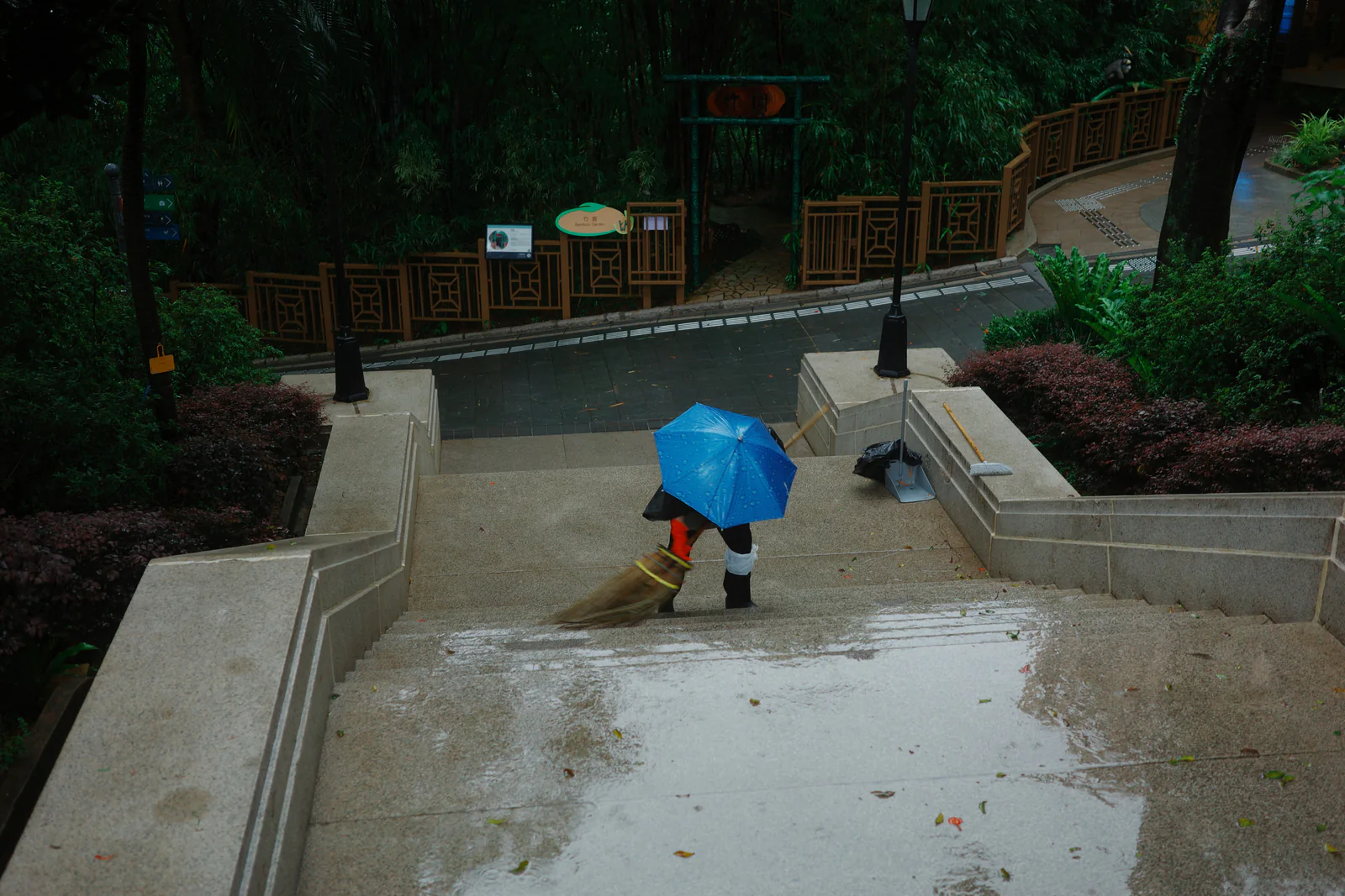 A worker sweeping rain-soaked steps beneath a bright blue umbrella