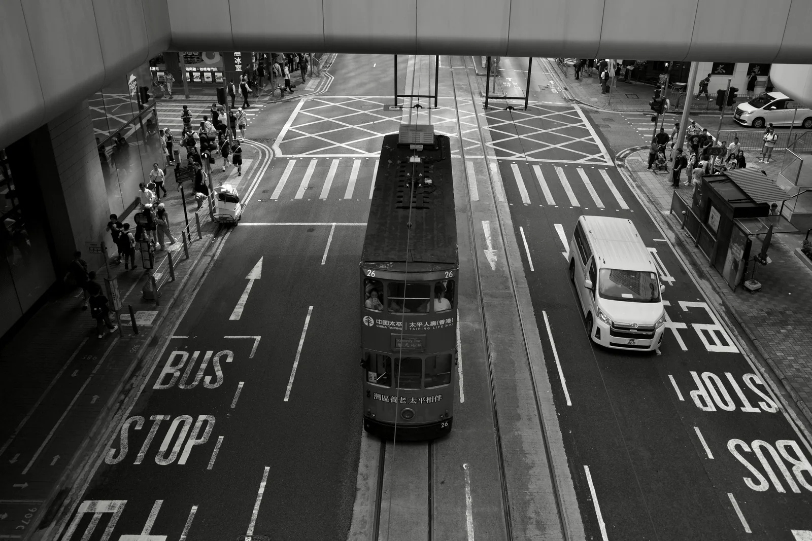 A double-decker tram and white van crossing a marked city intersection from above