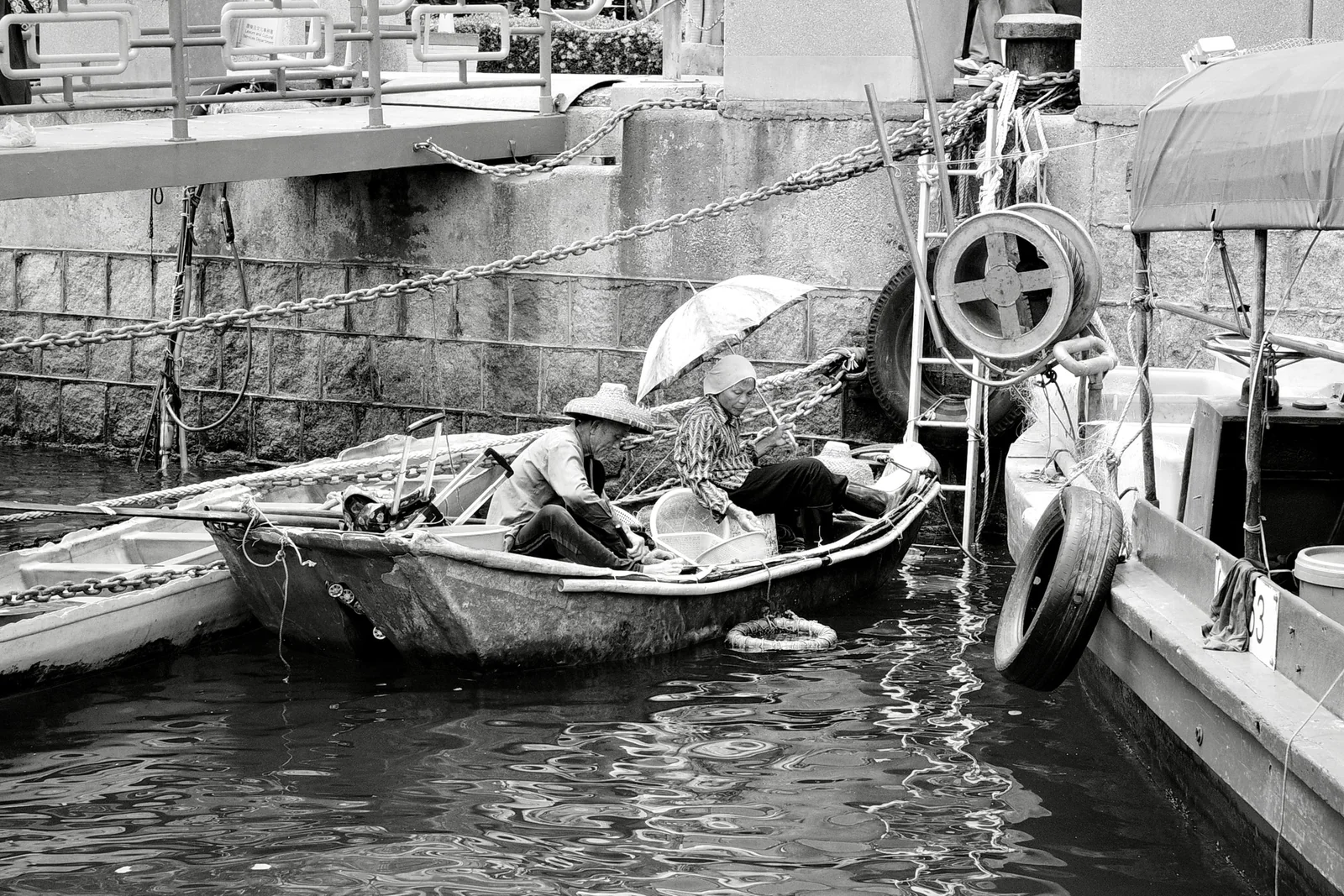 Two workers seated in a small harbor boat beneath umbrellas and heavy chains