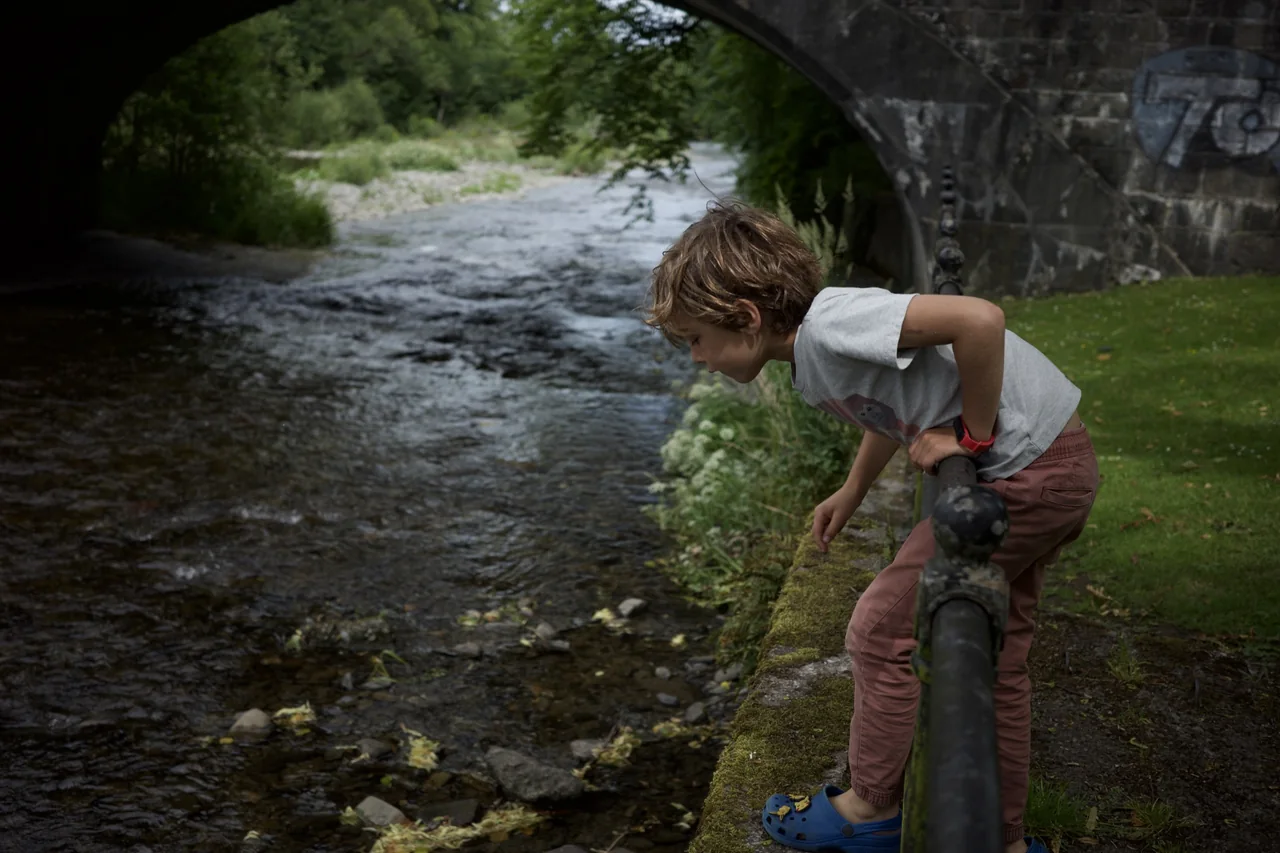 A child leaning over a railing to watch a stream beneath a stone bridge